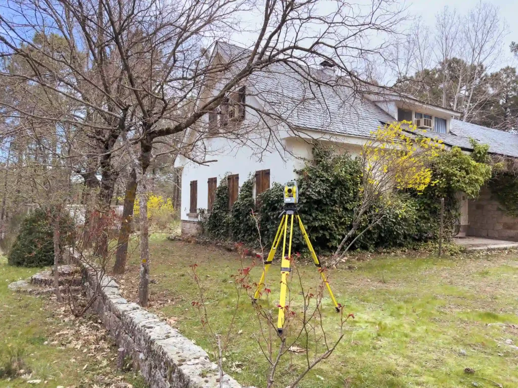 Una estación total amarilla montado sobre un trípode está ubicado en un jardín con césped frente a una casa de estilo rural.