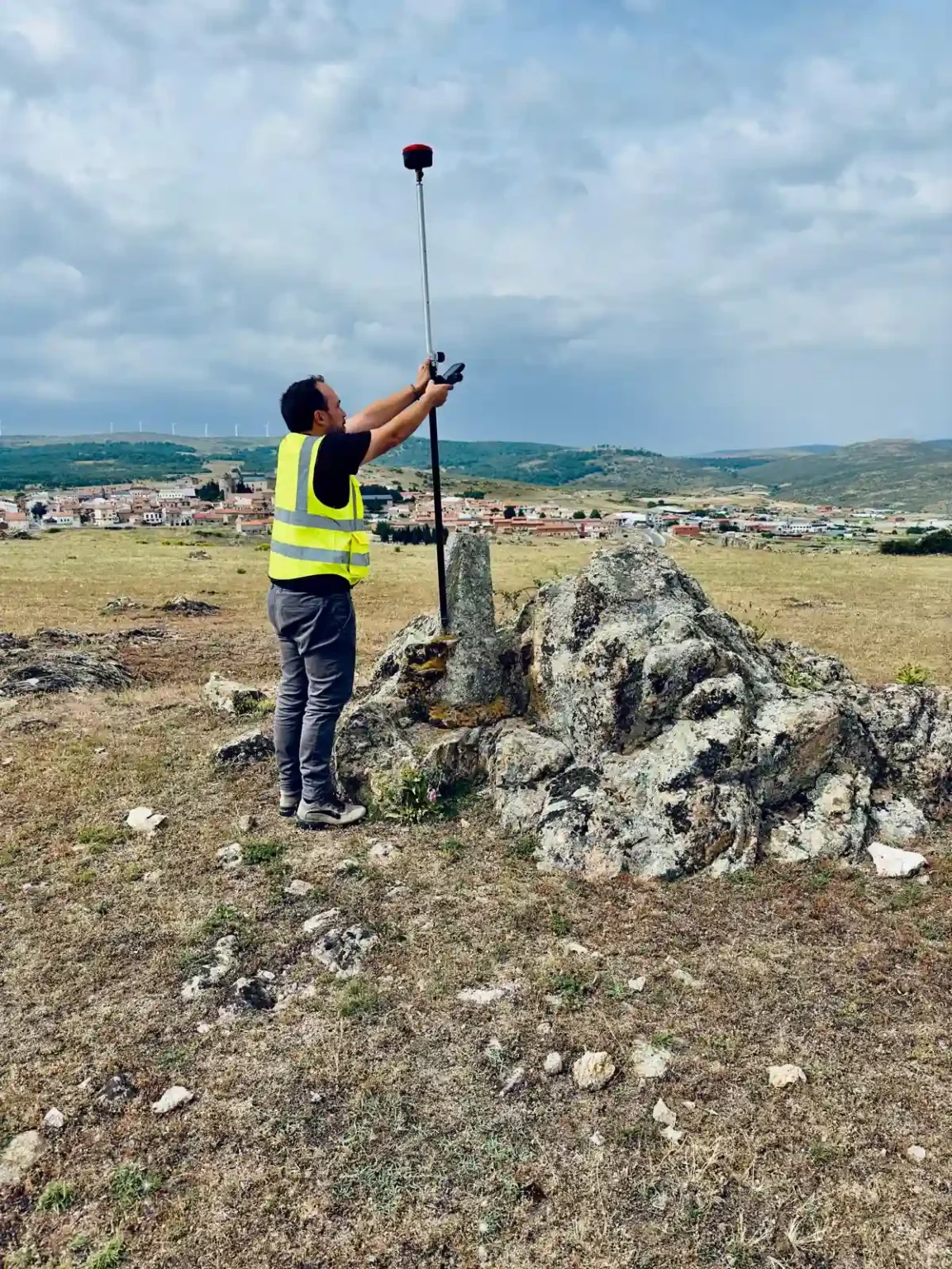 Un topógrafo con chaleco reflectante sostiene una vara con un receptor GNSS en la cima de una colina rocosa. Está tomando mediciones geoespaciales sobre un punto de referencia marcado por un mojón de piedra. En el fondo se observa un pueblo con casas y aerogeneradores en el horizonte, bajo un cielo parcialmente nublado.