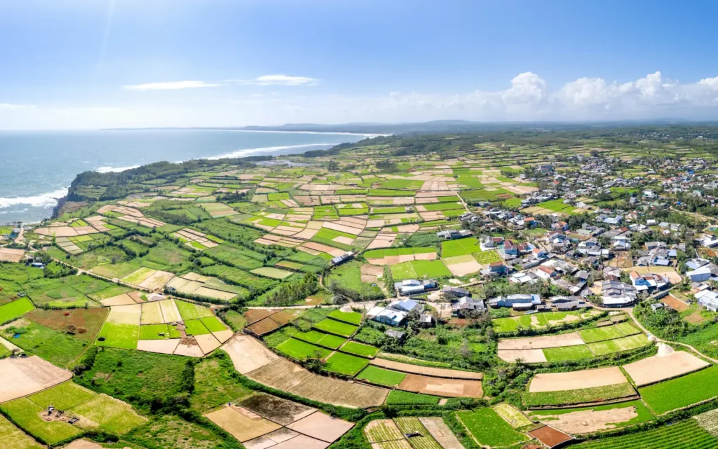 Imagen panorámica desde el aire de una zona rural con campos de cultivo de diversos tonos verdes y marrones, un pueblo con casas de techos metálicos y la costa con el océano en el horizonte.