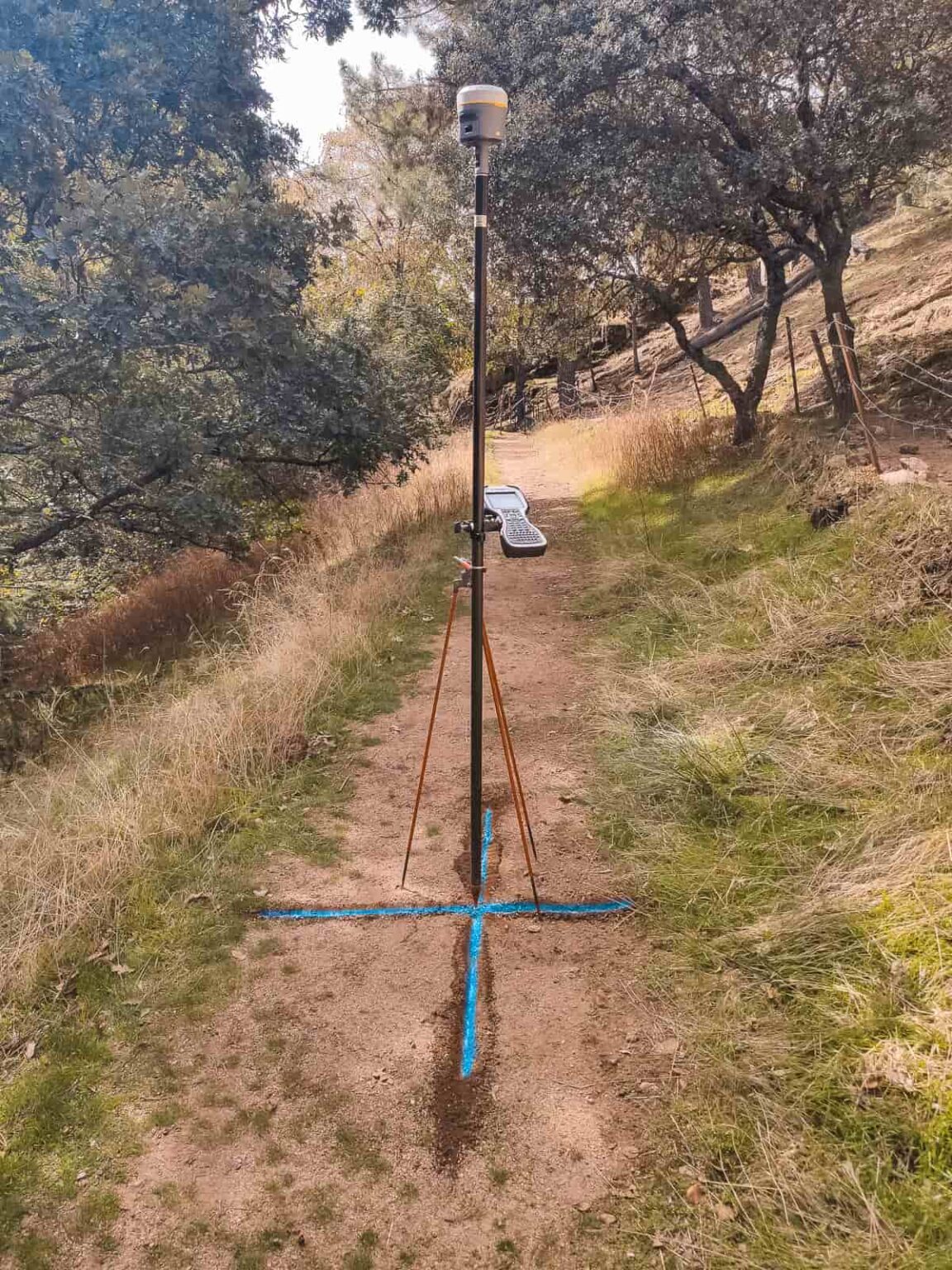 Un equipo de GPS geodésico GNSS montado en una vara con trípode se encuentra sobre un camino de tierra en una zona boscosa.